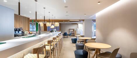 Un café moderne doté d’un long comptoir, d’un espace dédié aux baristas et d’un coin salon avec des tables et des chaises rondes.