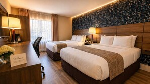 Une chambre d’hôtel avec deux lits, un bureau agrémenté d’une pancarte « Bienvenue », un miroir, une lampe et un mur orné d’un motif texturé.