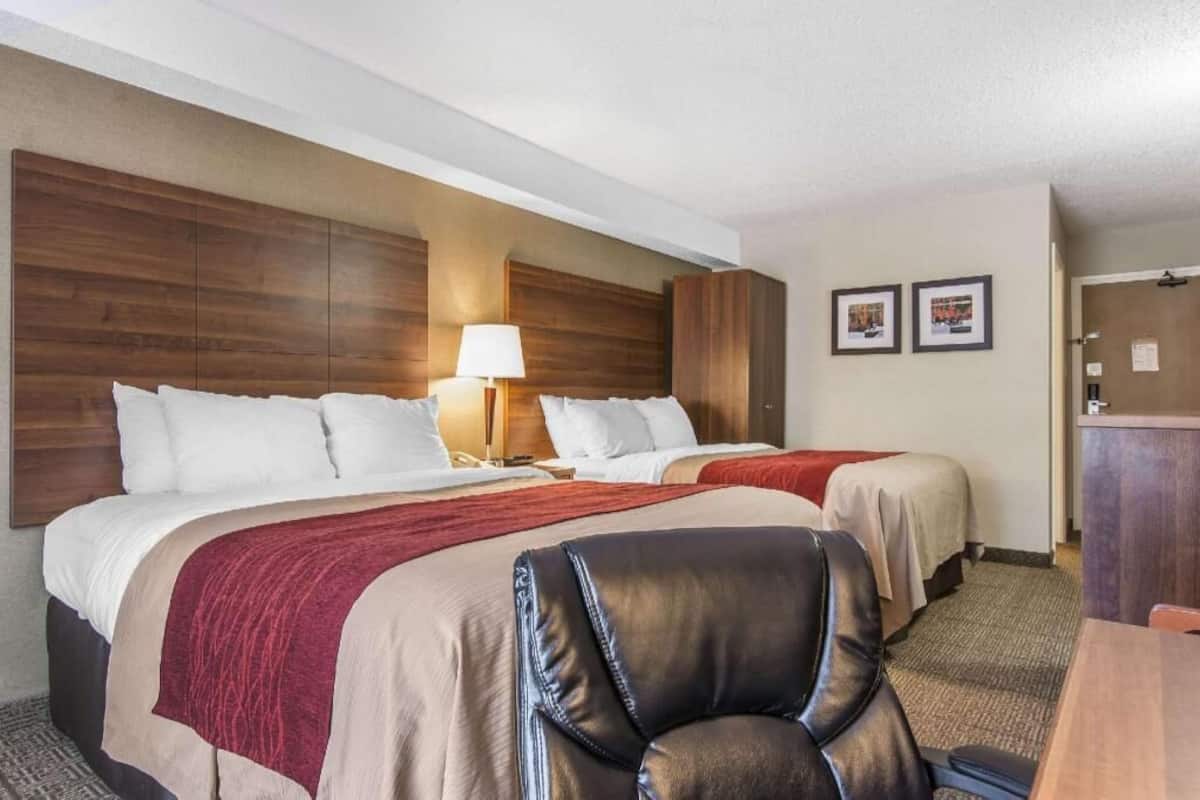 A hotel room with two beds, a desk, a chair, and a lamp.