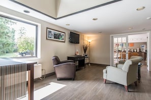 A modern living room with a large window, a flat-screen TV, a dining area, and a kitchen visible through an open door.