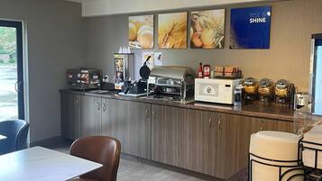 Uma cozinha moderna com máquina de waffle, cafeteira e micro-ondas. Há quadros nas paredes e uma placa com a inscrição 'RISE & SHINE'.