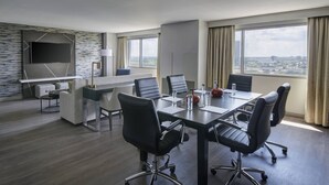 A modern hotel room with a large conference table, black office chairs, a flat-screen TV mounted on the wall, and large windows offering a city view. - Detroit Marriott Troy (Troy)