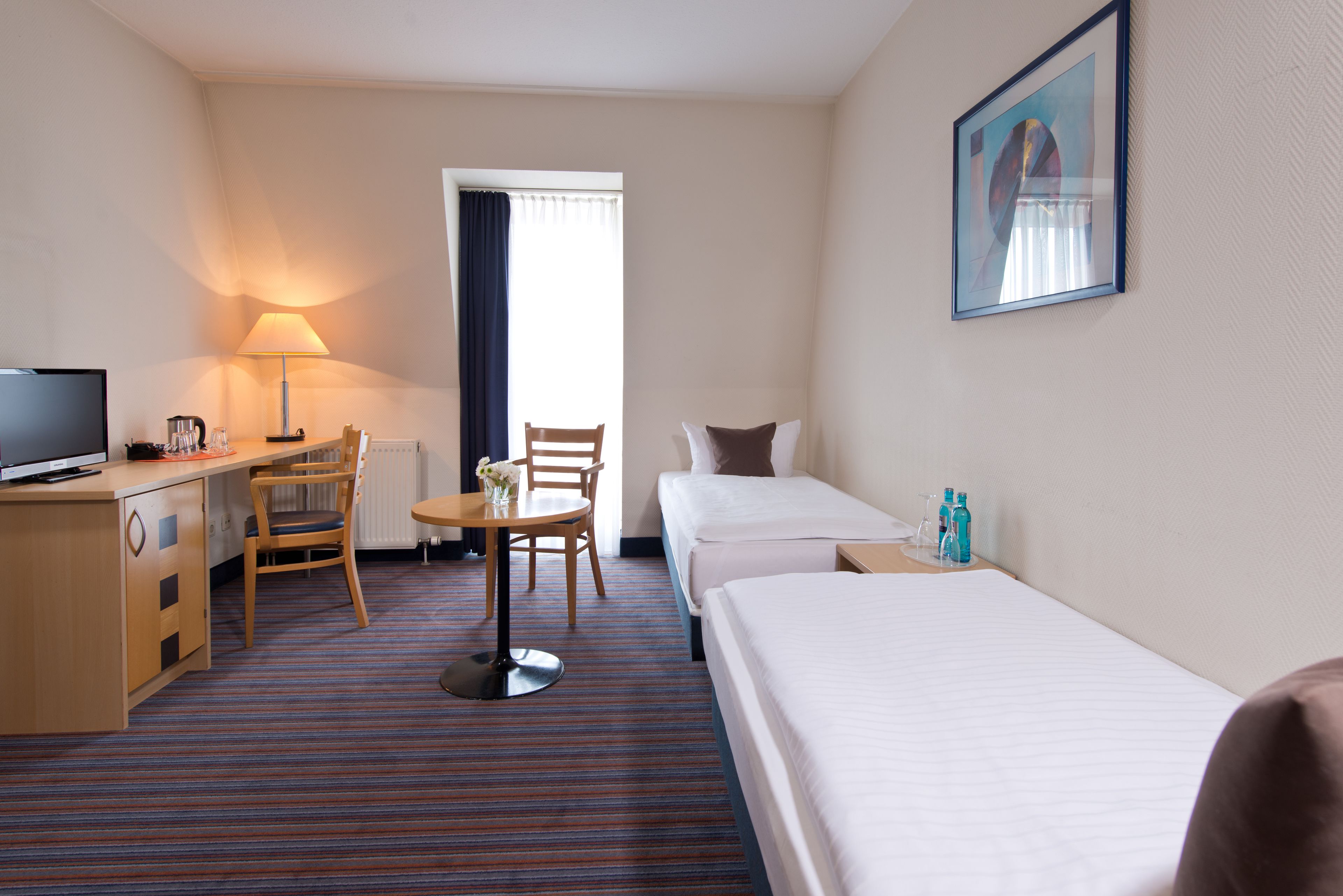 A hotel room with a bed, a desk with a lamp, a chair, a small table, and a television.
