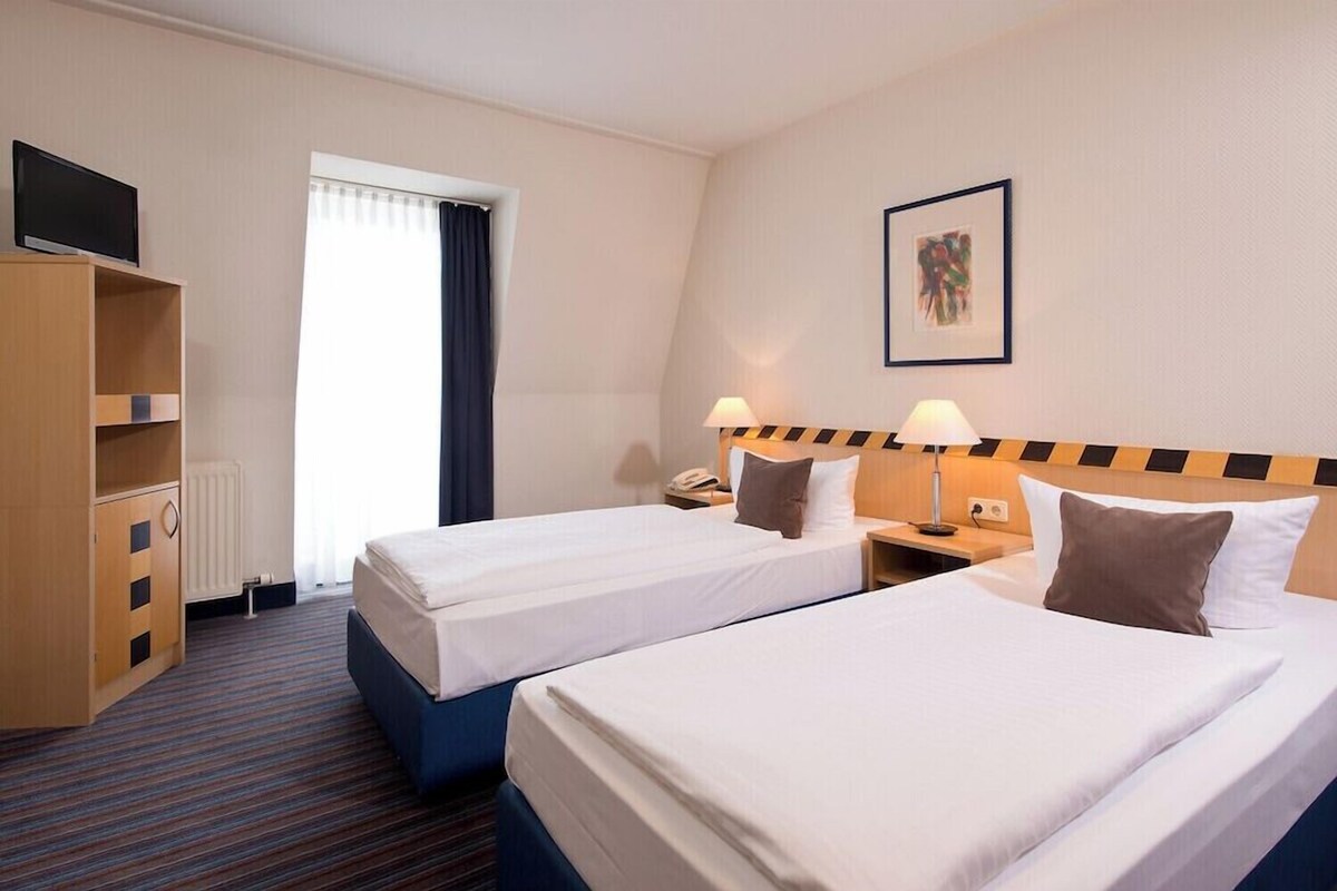 Habitación de hotel con dos camas, televisión, armario, escritorio con lámpara y ventana con cortinas.