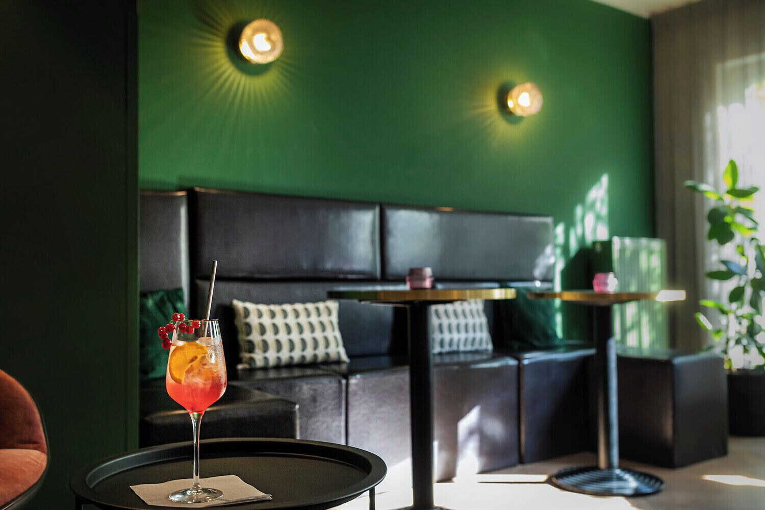 Un área de estar moderna con un sofá, una mesita redonda con una bebida y una pared verde con dos luces de pared.