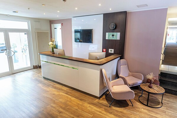 A hotel reception area with a reception desk, two cushioned chairs, a clock, and a small table with a vase.