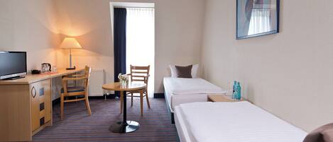 A hotel room with a bed, a desk with a TV, a chair, a small table with a vase, and a window with curtains.