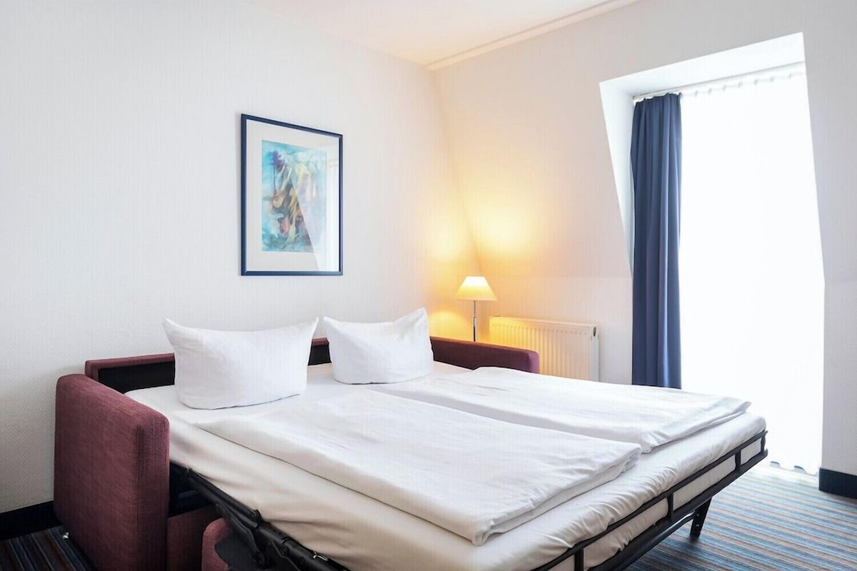 Habitación de hotel con cama doble, dos almohadas blancas, un sillón rojo, un cuadro en la pared, una lámpara de noche y una ventana con cortinas.
