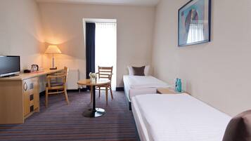 A hotel room with a bed, a desk with a TV, a chair, a small table with a vase, and a window with curtains.