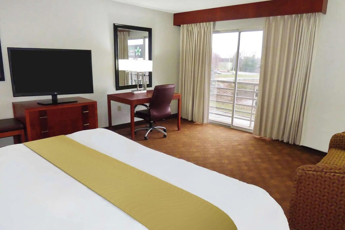 A hotel room with a bed, a desk with a chair, a television, a mirror, and a window with curtains.