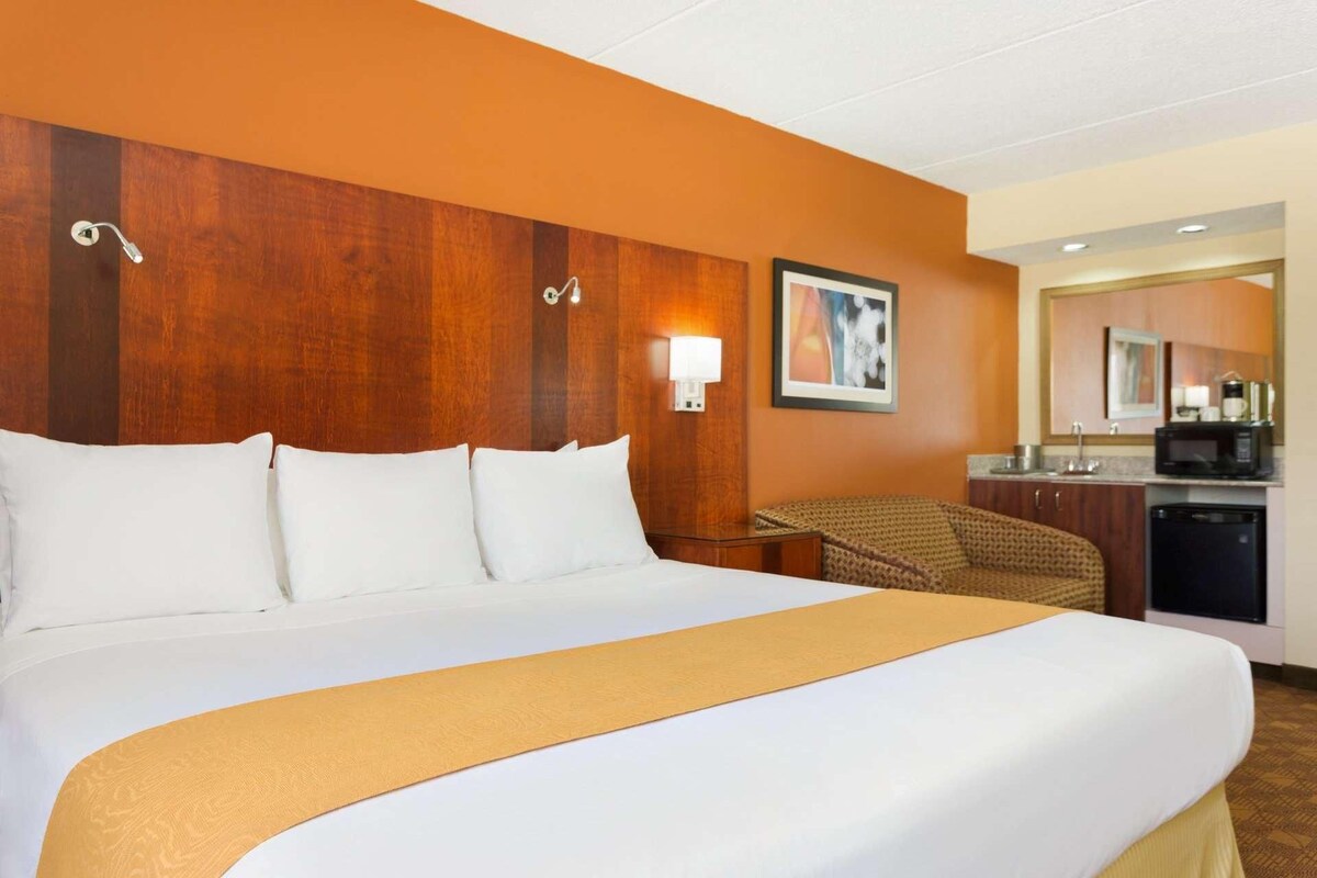 A hotel room with a large bed, a wooden headboard, a wall-mounted lamp, a small table, a chair, and a kitchenette in the background.