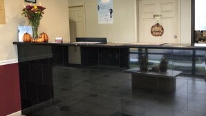 Un área de recepción con un mostrador negro, un tapete de bienvenida, un certificado enmarcado, un cartel y una puerta con la leyenda 