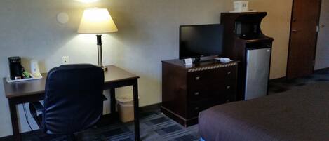 A hotel room with a desk, chair, lamp, television, minibar, and a bed.