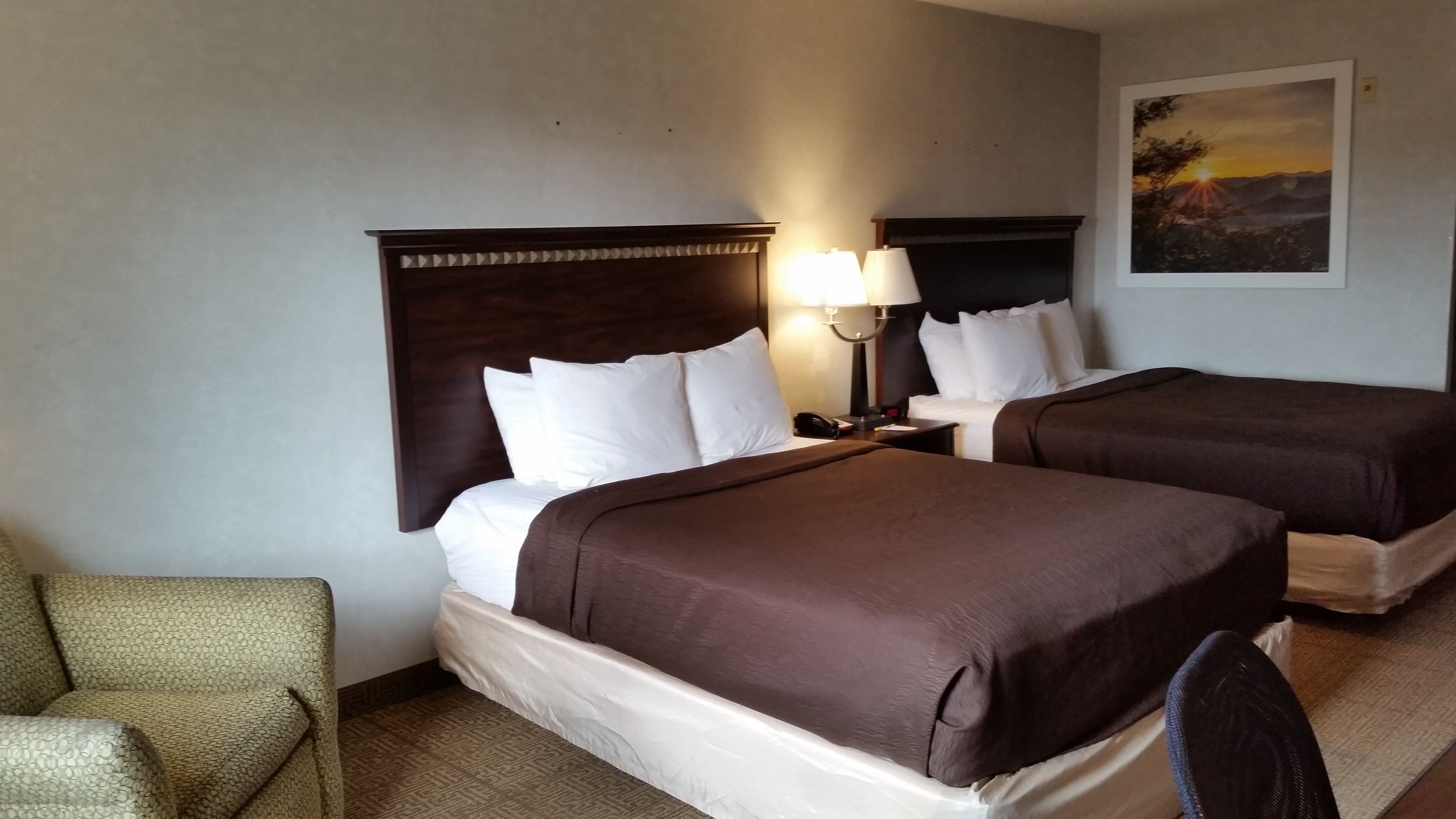 A hotel room with two beds, a nightstand with a lamp, a chair, and a painting on the wall.