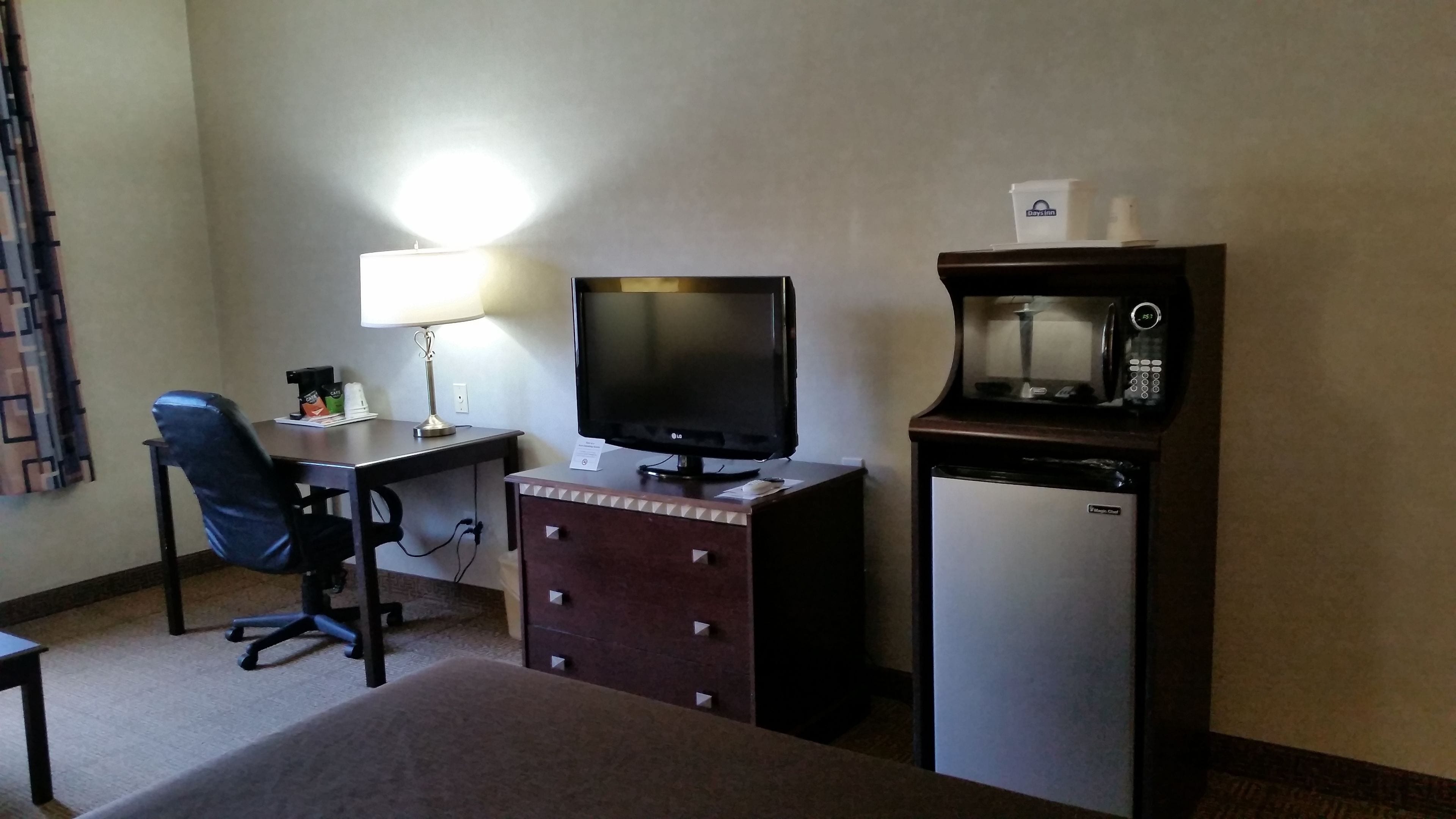 A hotel room with a desk, chair, lamp, television, microwave, and refrigerator.