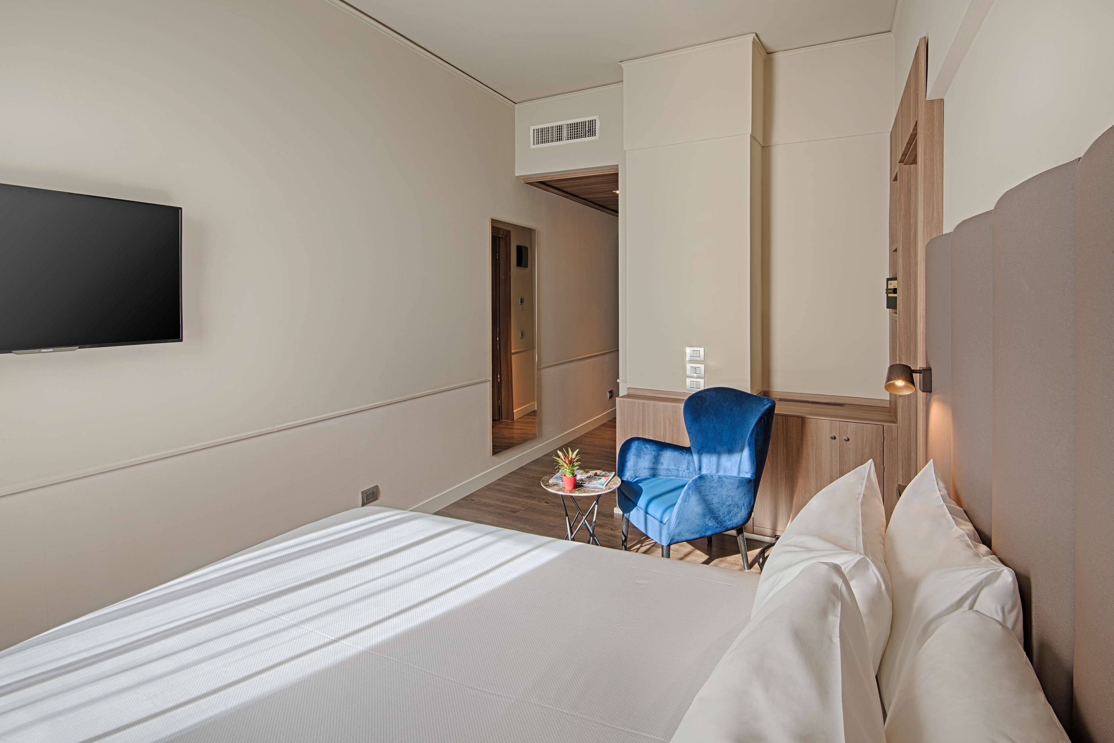 A hotel room with a bed, a blue armchair, a small table with a plant, and a TV mounted on the wall.
