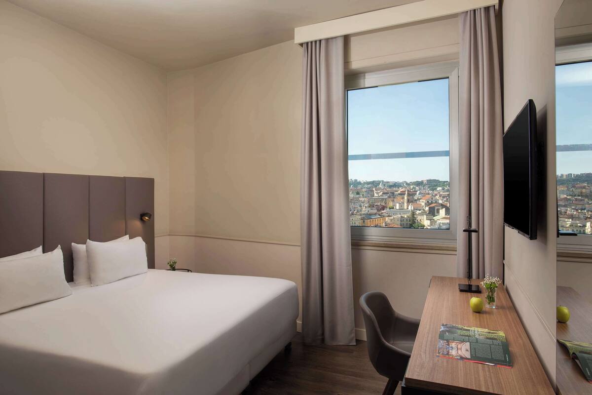A hotel room with a large bed, a desk with a chair, a flat-screen TV, and a window with a city view.