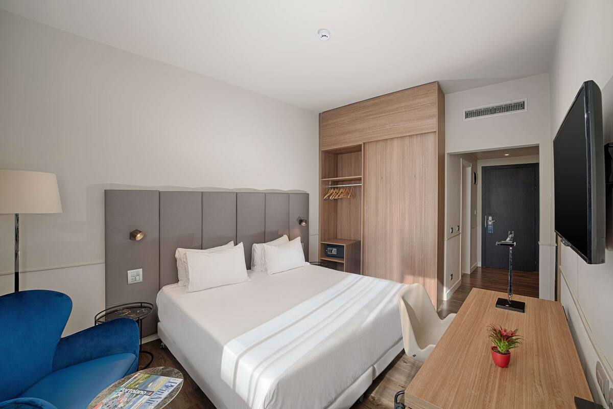 A hotel room with a large bed, a desk with a plant, a blue chair, and a TV.