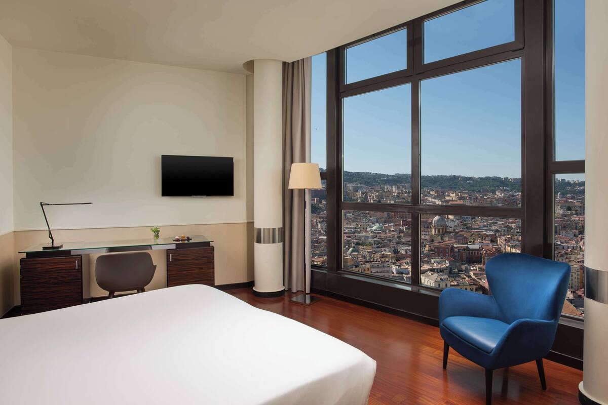 A hotel room with a large bed, a desk with a chair, a TV on the wall, and a view of a cityscape through large windows.