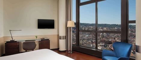 Habitación de hotel con una cama grande, un escritorio con silla, televisor en la pared y vistas a un paisaje urbano a través de amplios ventanales.