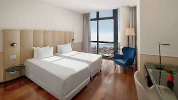 A hotel room with two beds, a desk, a chair, a view of the city, and a large window.