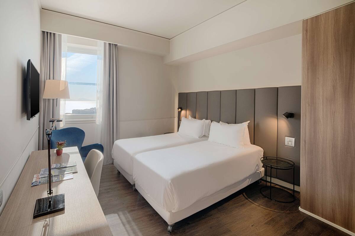 A hotel room with a large bed, a desk with a lamp, a chair, and a TV on the wall.