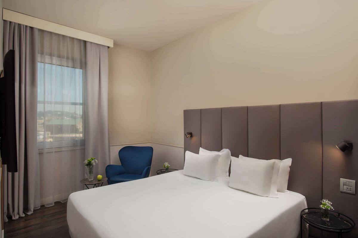 A hotel room with a large bed, a blue chair, a small table with a vase, and a view of the city through the window.
