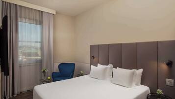 Ett hotellrum med en stor säng, en blå stol, ett litet bord med en vas och utsikt över staden genom fönstret.