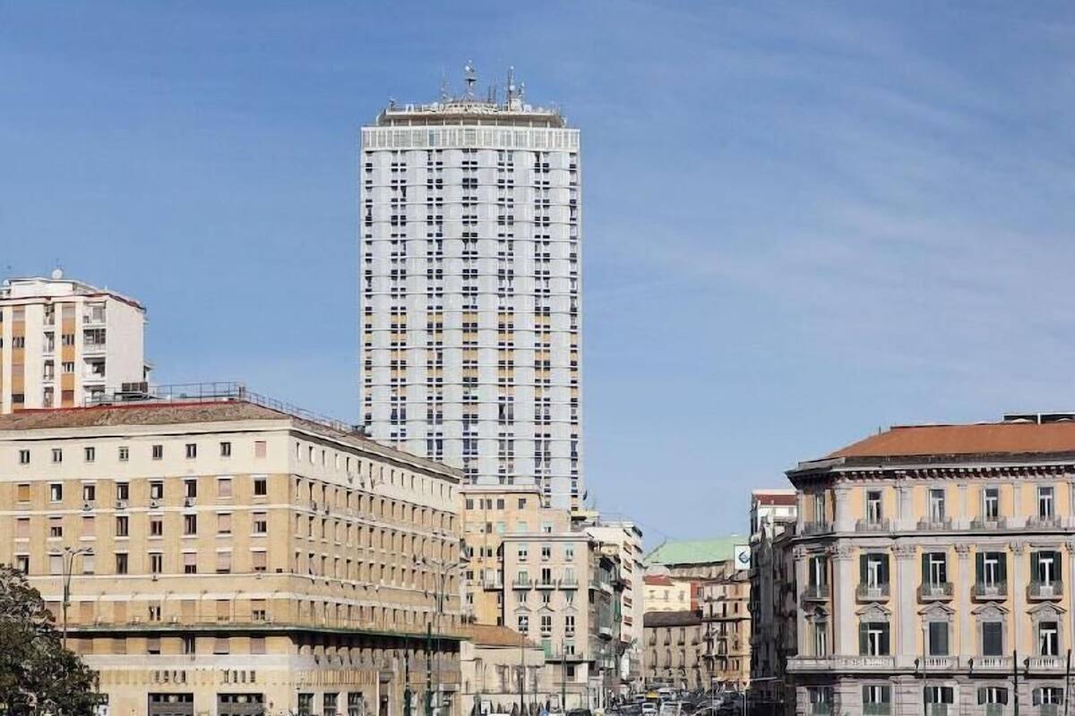 A tall, modern high-rise building with a grid of windows, surrounded by older, multi-story buildings with varied architectural styles.