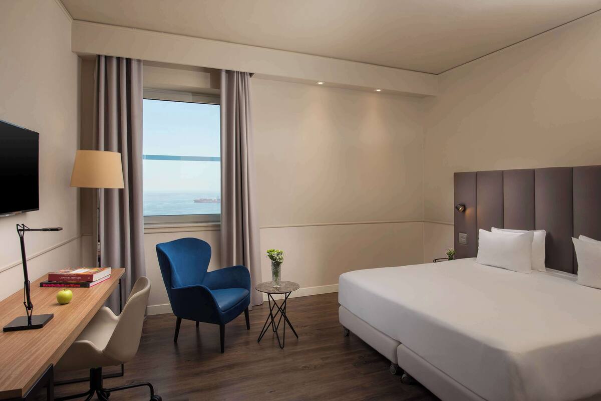 A hotel room with a bed, a desk with a lamp, a chair, a small table with a vase, and a view of the sea.