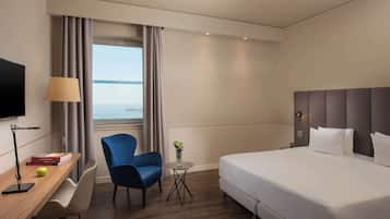 Ein Hotelzimmer mit einem Bett, einem Schreibtisch mit Lampe, einem Stuhl, einem kleinen Tisch mit Blumenvase und Blick aufs Meer.
