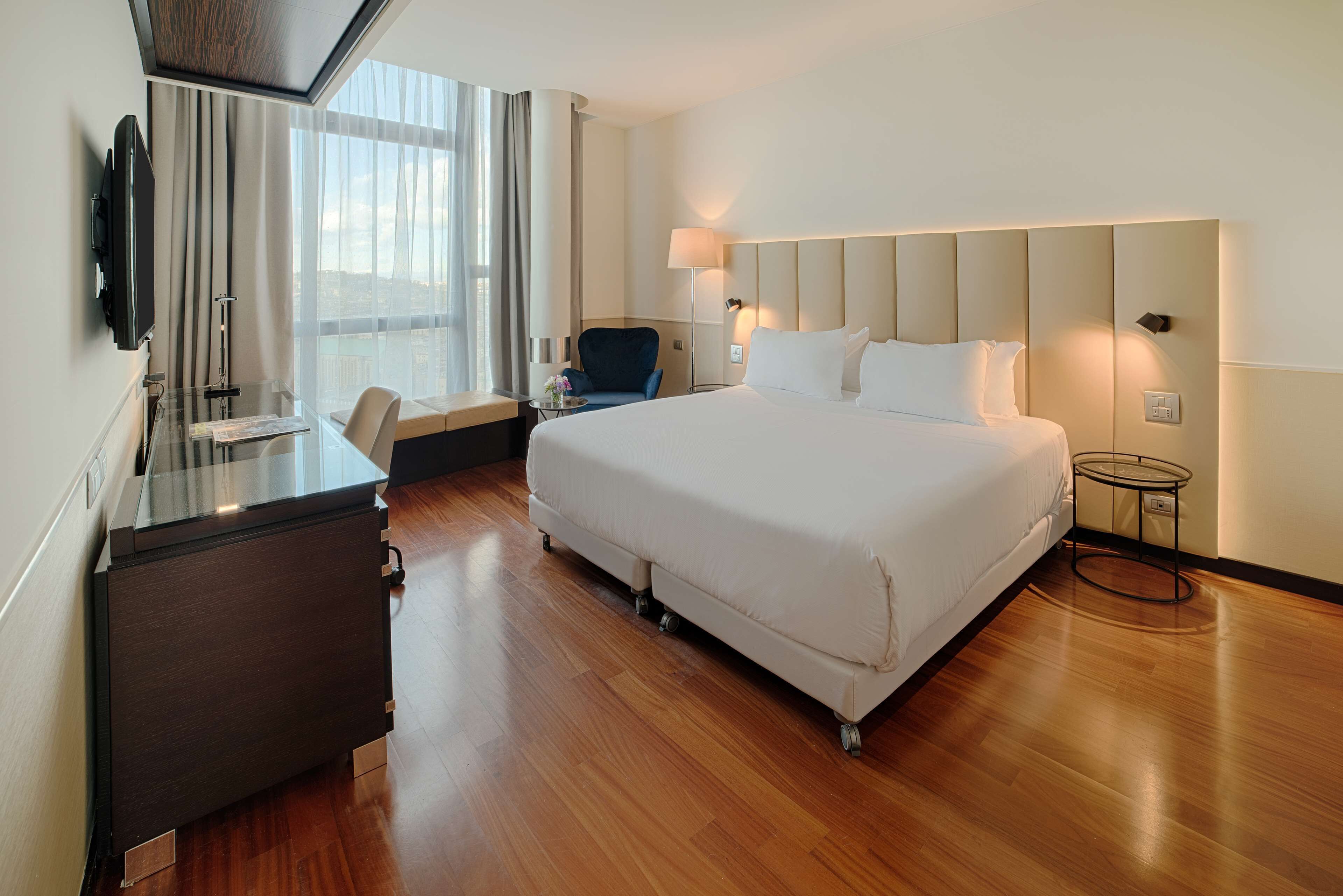 A hotel room with a large bed, a desk with a flat-screen TV, a chair, and a small table.