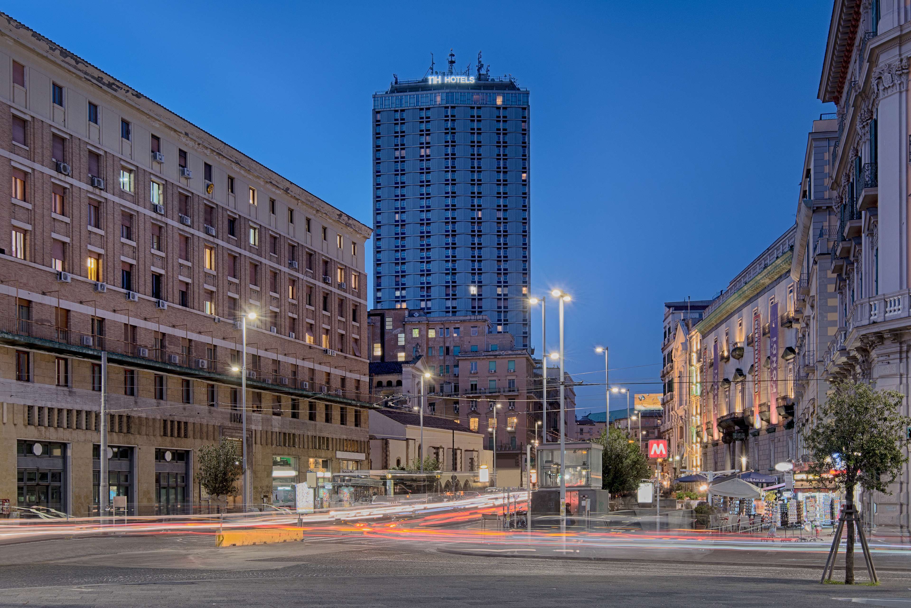 Ein Hochhaus-Hotel mit mehreren Etagen, einem gut sichtbaren 'TH HOTELS'-Schild und einer belebten Straße mit Lichtspuren, die den Verkehr anzeigen.