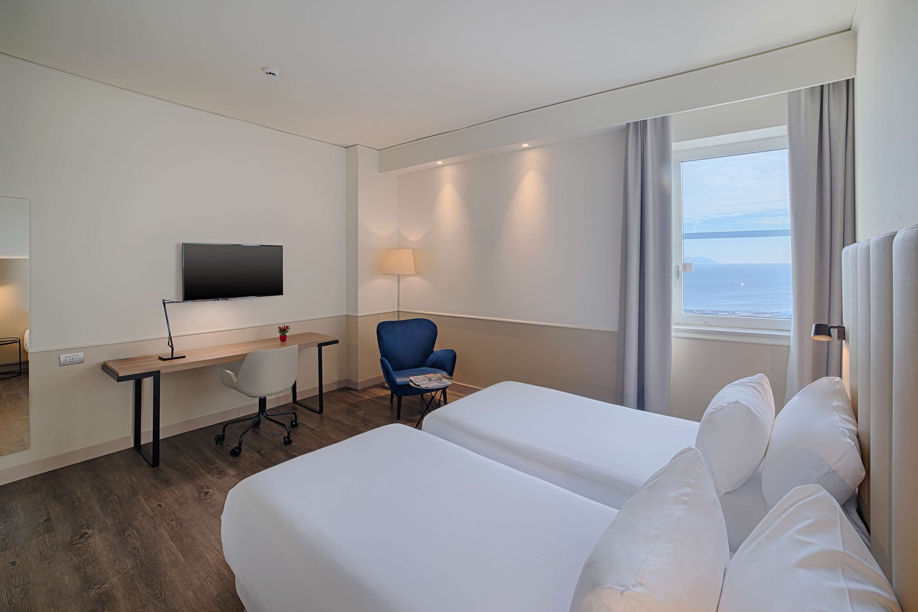 A hotel room with two beds, a desk, a chair, a TV, and a window with a view of the sea.