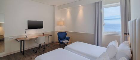 Habitación de hotel con dos camas, un escritorio, una silla, un televisor y ventana con vista al mar.