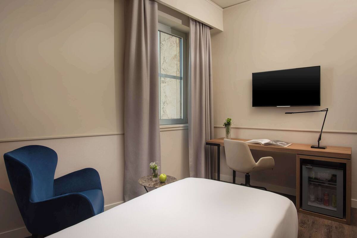 A hotel room with a bed, a desk with a television, a chair, a small table with a vase and an apple, and a mini-fridge.
