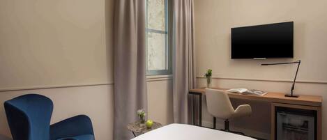 Une chambre d’hôtel comprenant un lit, un bureau avec une télévision, une chaise, une petite table avec un vase et une pomme, et un mini-réfrigérateur.