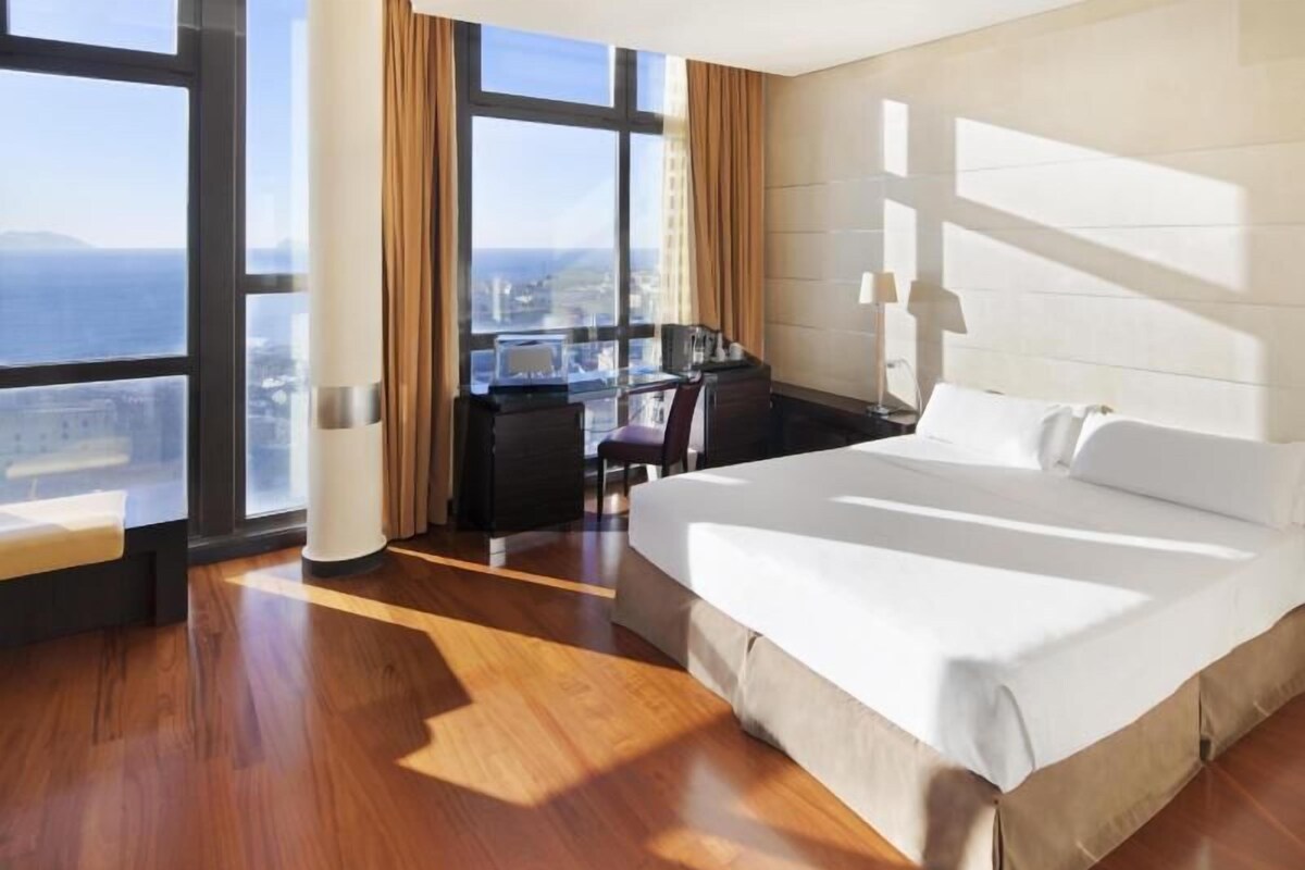 A hotel room with a large bed, a desk, a chair, and a view of the city through large windows.