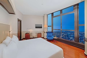 Una camera d'albergo con un letto grande, una scrivania, una sedia, una TV e un'ampia finestra vista città.