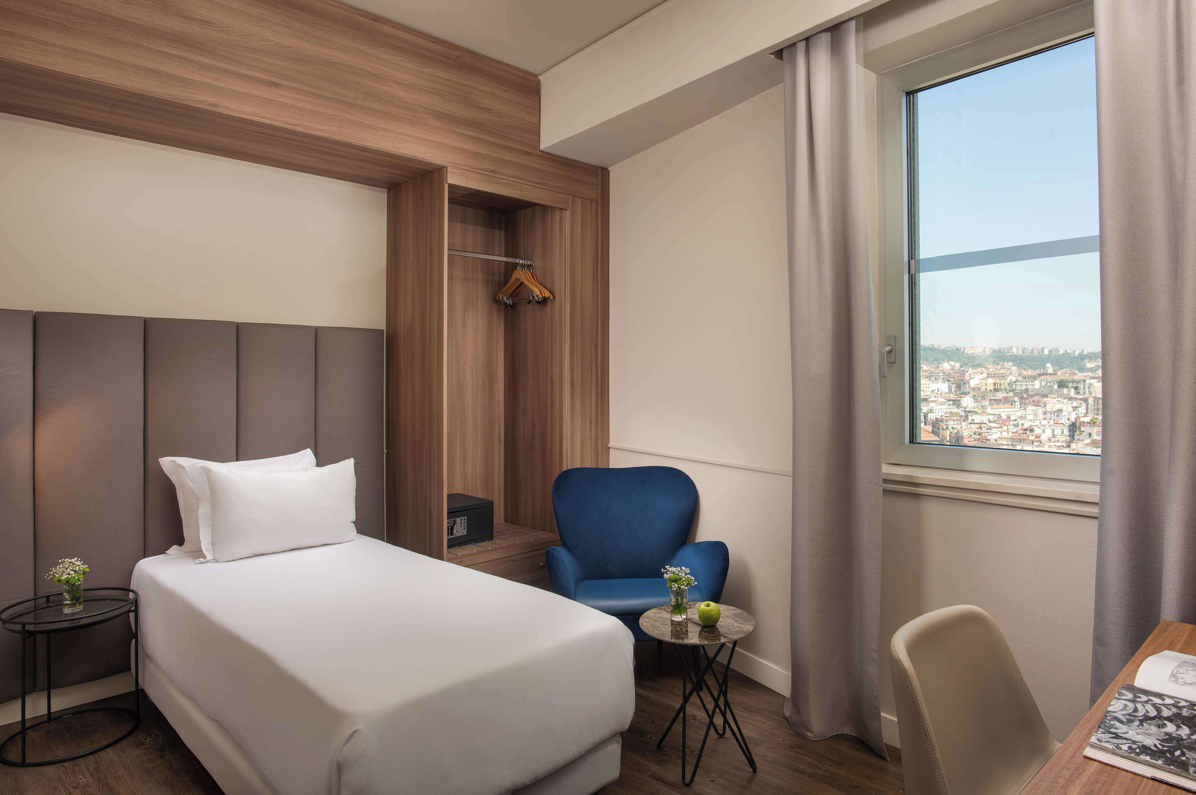 Habitación de hotel con cama, un sillón azul, una mesita con un jarrón y vistas a la ciudad a través de una ventana.