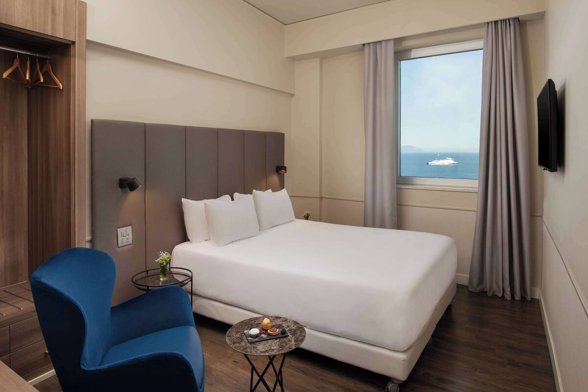 A hotel room with a large bed, a blue armchair, a small table with a tray of pastries, a TV mounted on the wall, and a window with a view of the sea.