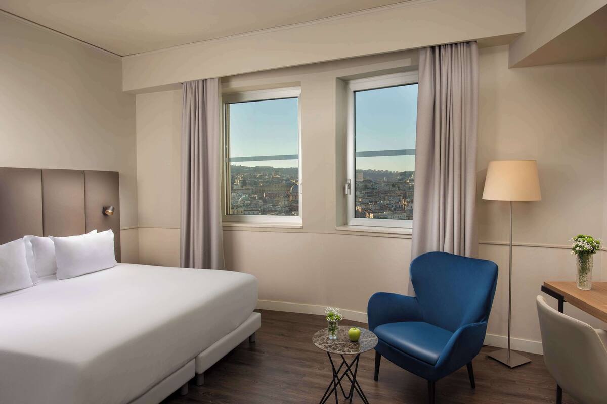 A hotel room with a large bed, a blue armchair, a small table with a lamp, and a view of a cityscape through two windows.