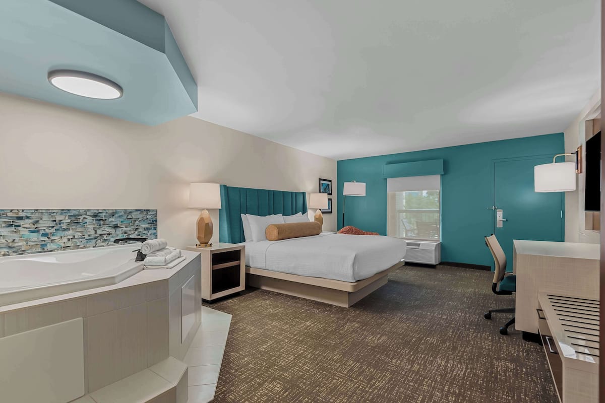 A modern hotel room with a large bathtub, a bed with teal headboard, a desk, and a window with a view.
