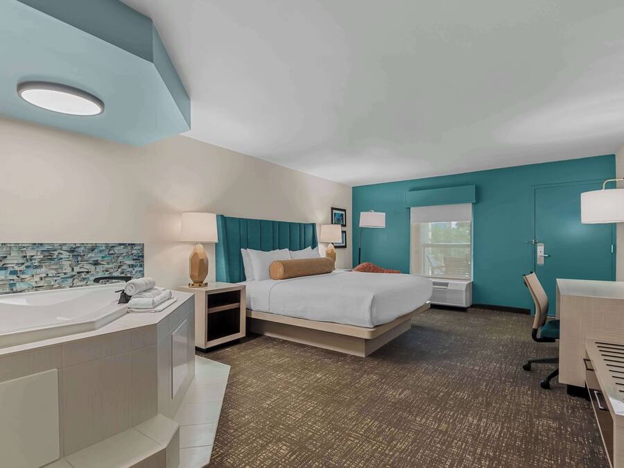 A modern hotel room with a large bathtub, a bed with teal headboard, a desk, and a window with a view.
