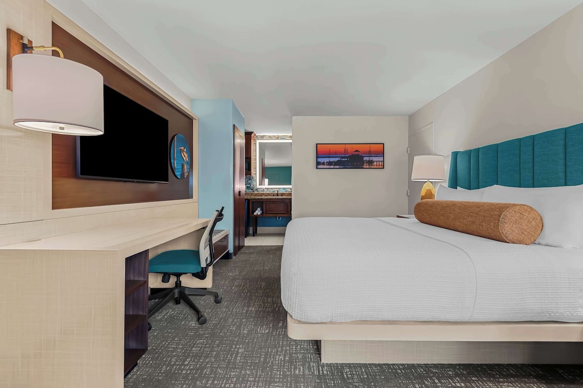 A hotel room with a bed, desk, chair, TV, artwork, and a lamp.