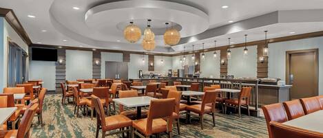 Une salle à manger d’hôtel avec des tables rondes et des chaises orange, une cuisine en arrière-plan et un tapis à motifs.