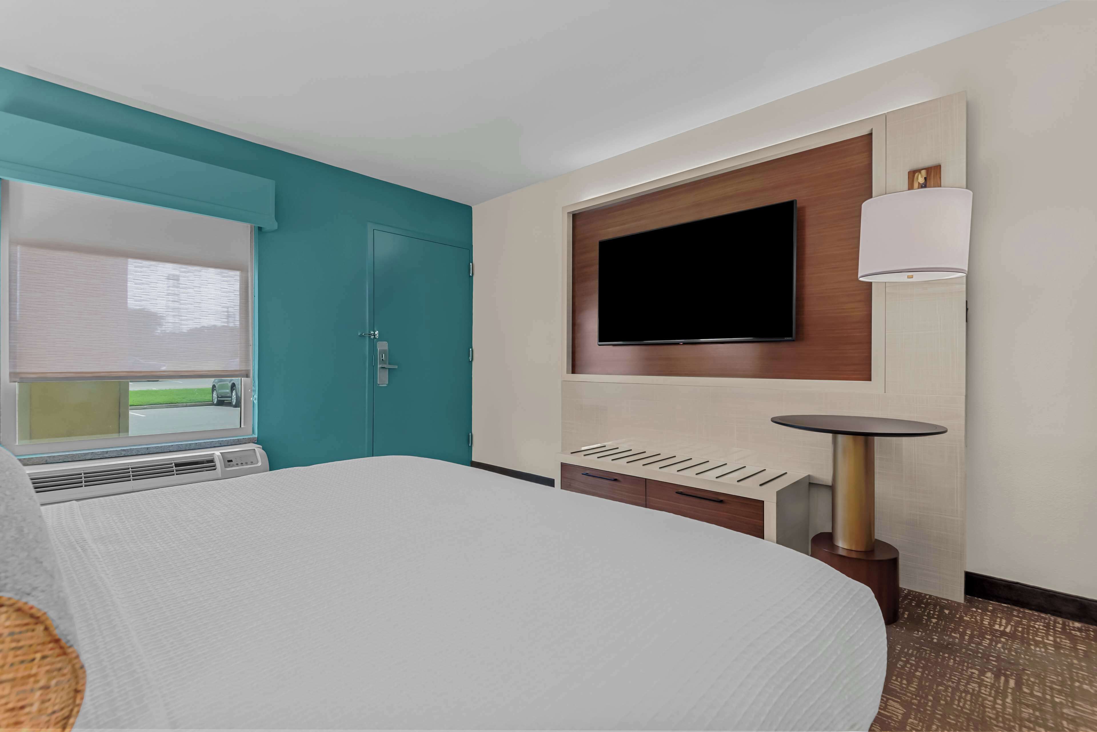 Een hotelkamer met een bed, een aan de muur bevestigde televisie, een klein tafeltje en een raam met jaloezieën.