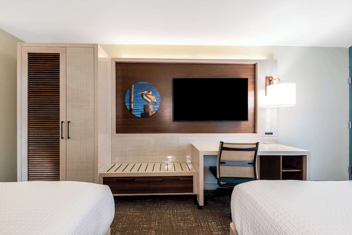 A hotel room with two beds, a desk, a chair, a television, and a wall-mounted lamp.
