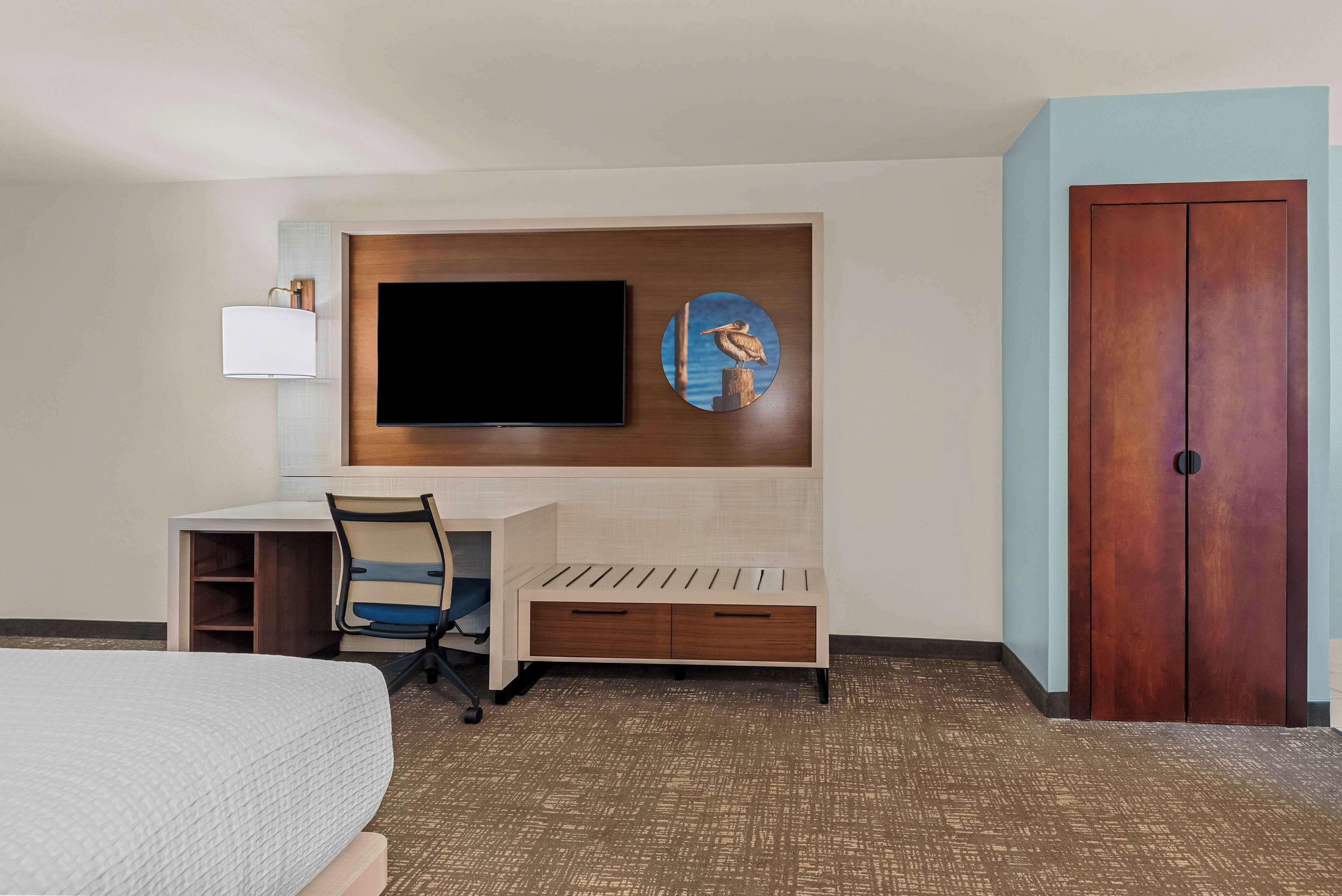Habitación de hotel con una cama, un escritorio con silla, un televisor montado en la pared, un armario de madera y una obra de arte circular.