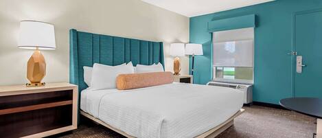 A hotel room with a large bed, two bedside tables, a window with blinds, and a teal accent wall.